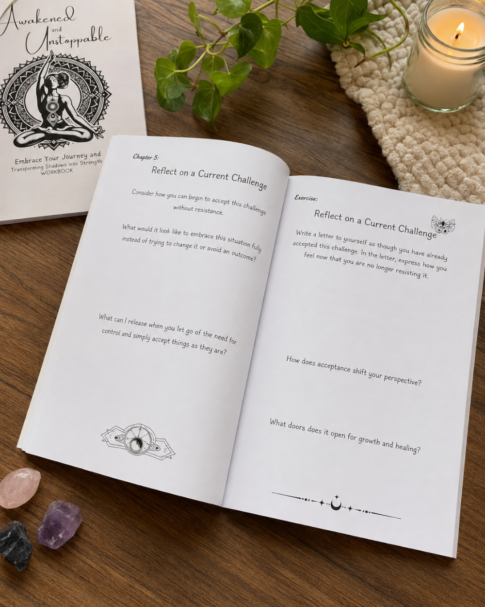 Awakened and Unstoppable workbook by Wilmaris Santos spread on a wooden table, showing reflective prompts from Chapter 5 'Reflect on a Current Challenge.' A second closed copy of the journal is visible in the upper left, along with a lit candle in a glass jar, a knitted coaster, and small crystals on the tabletop.