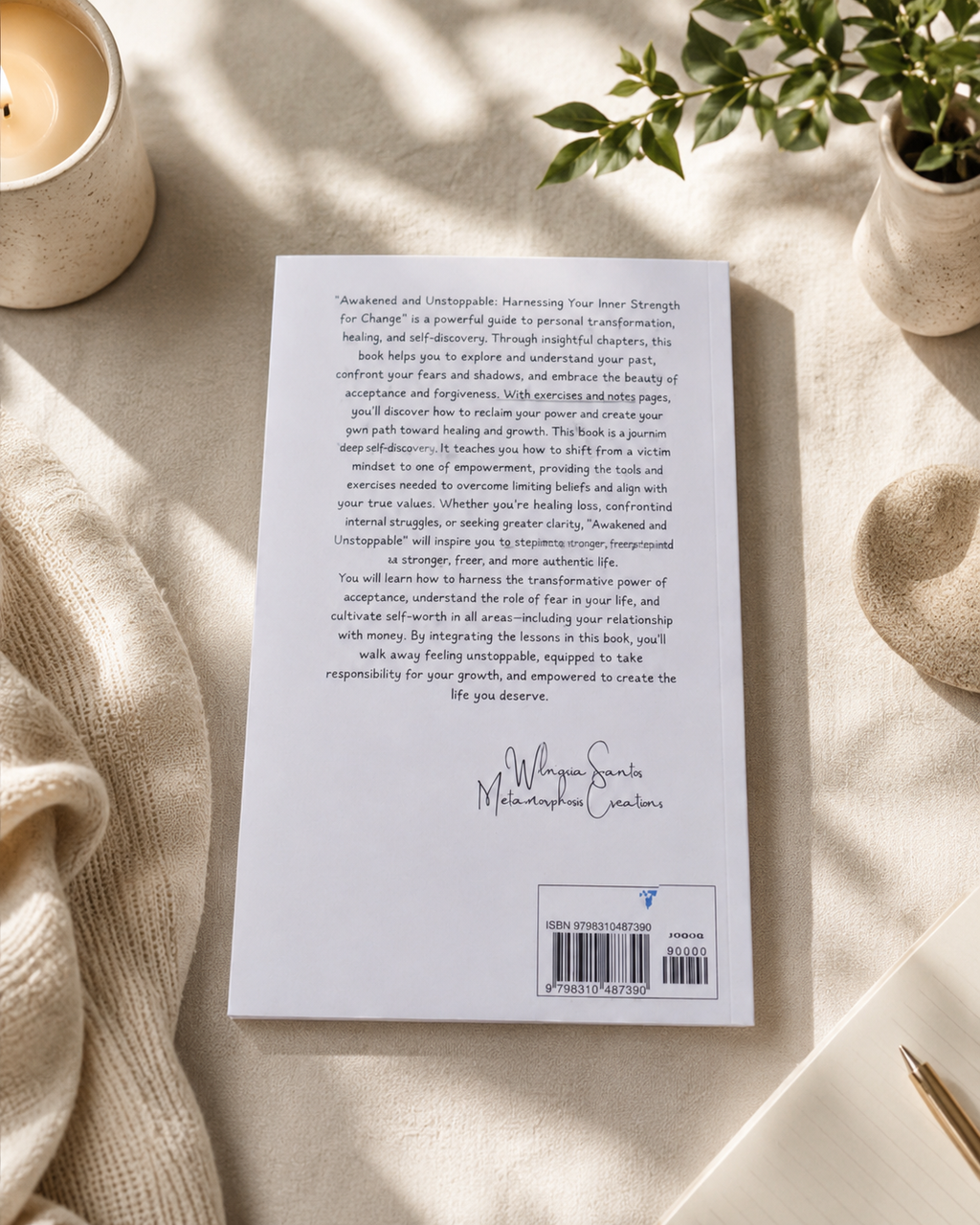 The back cover of Awakened and Unstoppable paperback workbook by Wilmaris Santos is displayed on a beige textured blanket, styled with a lit candle, a small potted plant, and a gold pen.