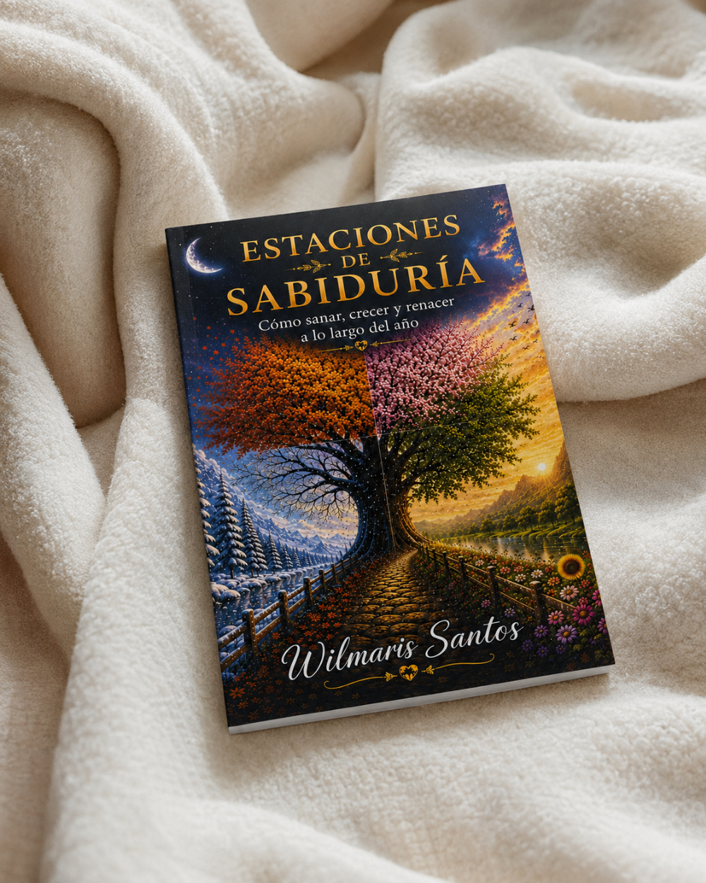 A book titled 'Estaciones de Sabiduría' by Wilmaris Santos lies on a soft, cream-colored fleece blanket. The cover features a colorful illustration of a tree with seasonal foliage (blue, pink, green, orange) along a path, under a crescent moon.