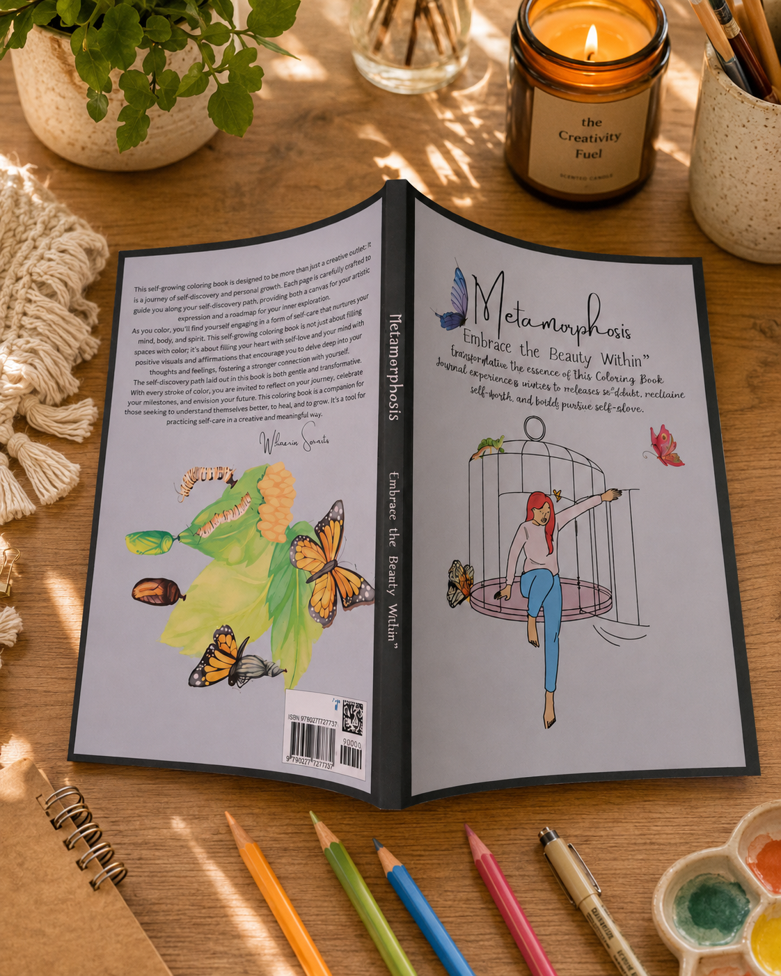 An open hardcover book titled 'Metamorphosis Embrace The Beauty Within' is displayed on a wooden desk, showing both the front and back covers. The book is surrounded by creative and study props, including colored pencils, a paint palette, a lit candle in a ceramic jar, and a potted plant in the background.