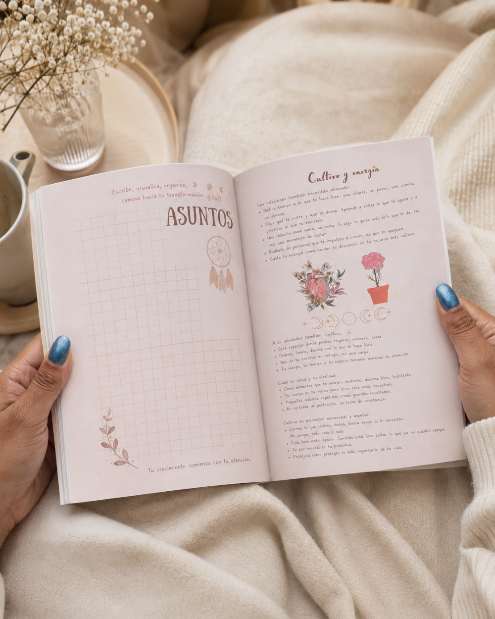 Transformandome by Wilmaris Santos spread held by two hands with blue nail polish, resting on a soft beige blanket. The pages are cream-colored with Spanish text, grid lines, and delicate floral illustrations.