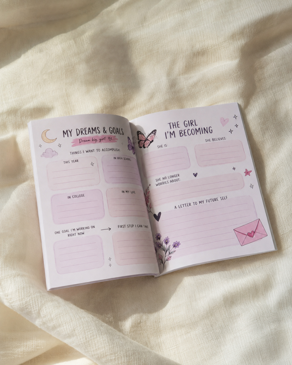 An open pastel planner or journal spread on soft cream fabric, with handwritten sections titled 'My Dreams & Goals' and 'The Girl I'm Becoming' decorated with small pink and purple illustrations.