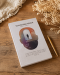 A white notebook titled 'Transformandome by Wilmaris Santos' with a colorful graphic on the cover, styled on a wooden table with dried flowers, a beige cloth, and a gold pen.